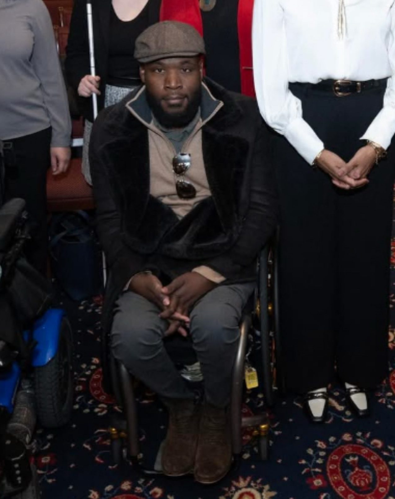 'n Man met 'n plat pet, donker jas en stewels sit in 'n rolstoel, met sy hande saamgevou. Hy is omring deur mense, sommige staan, op 'n patroonmattevloer.